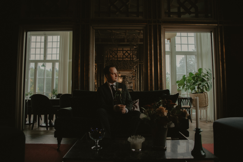 Brudgum i mörk kostym sitter i Villa Strömsfors eleganta salong, omgiven av ljus från stora fönster, ett porträtt fyllt av lugn och förväntan.